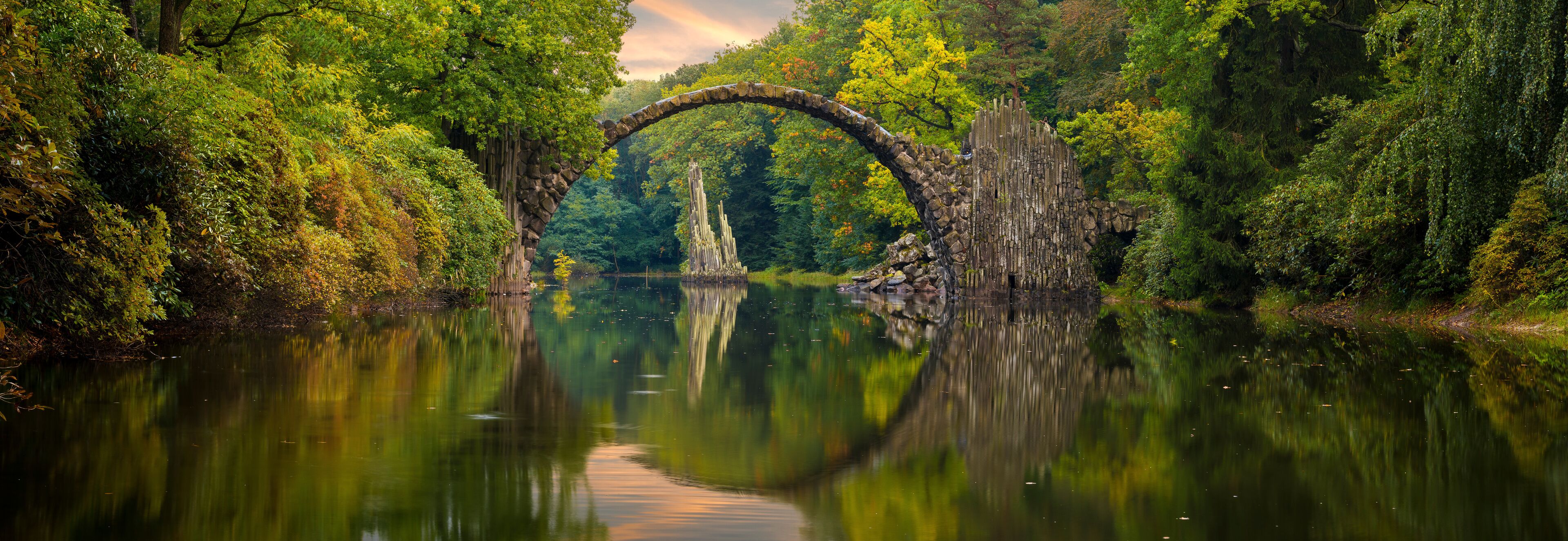 A stone arch bridge over a calm river surrounded by lush greenery.
