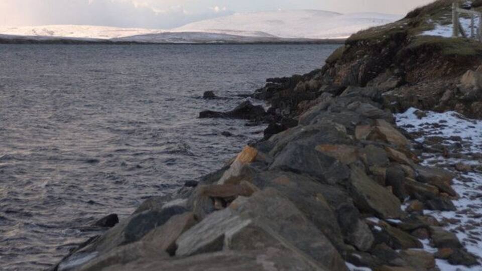 A rocky shoreline with snow patches and a distant mountain range.