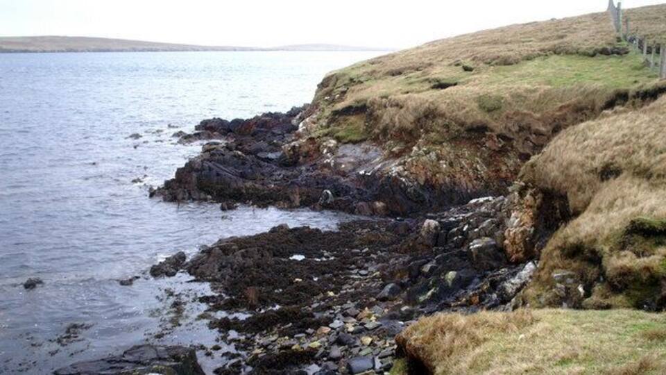 A rocky coastline with a grassy hill and a distant structure.