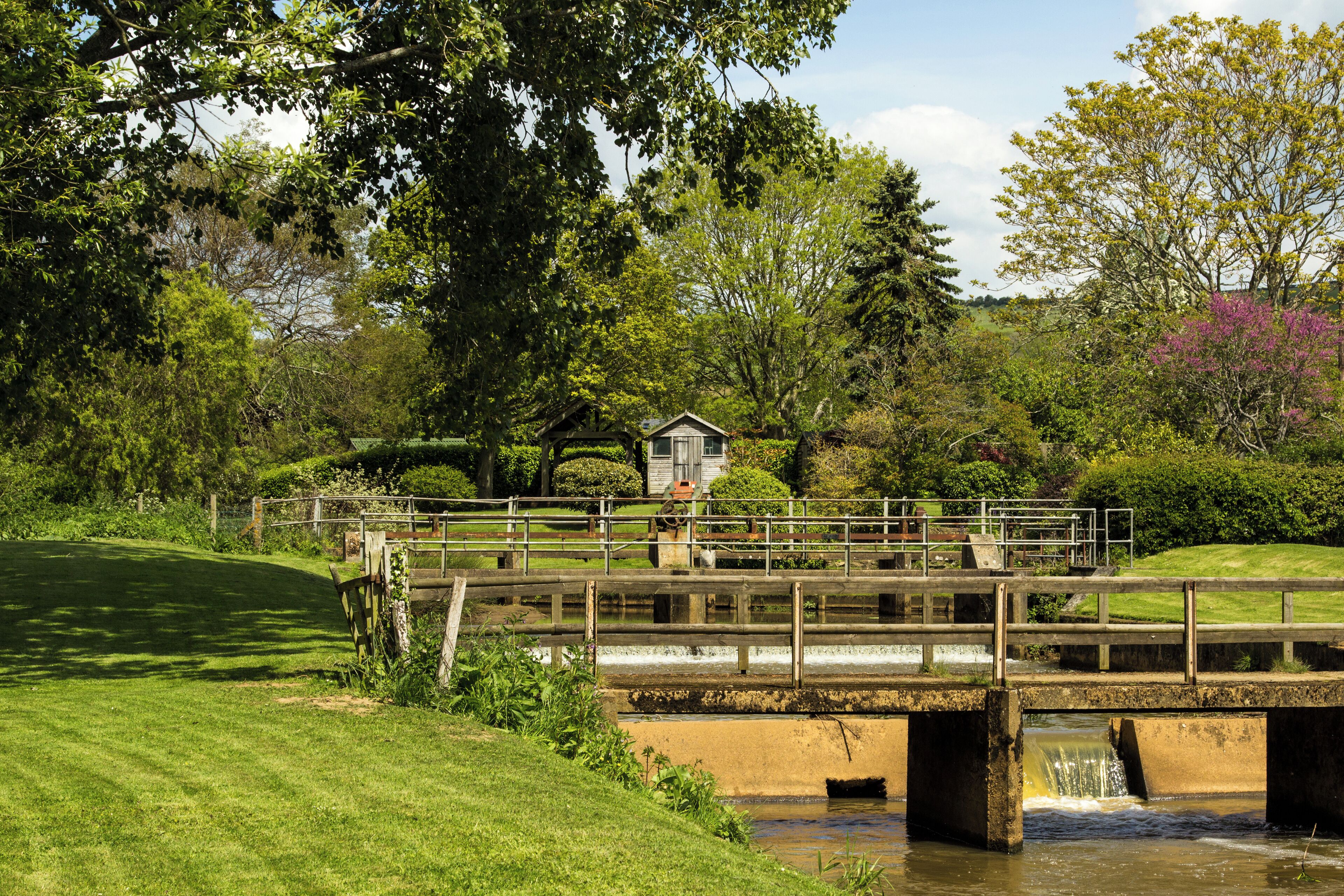 A wooden bridge over a small stream with a house in the background.