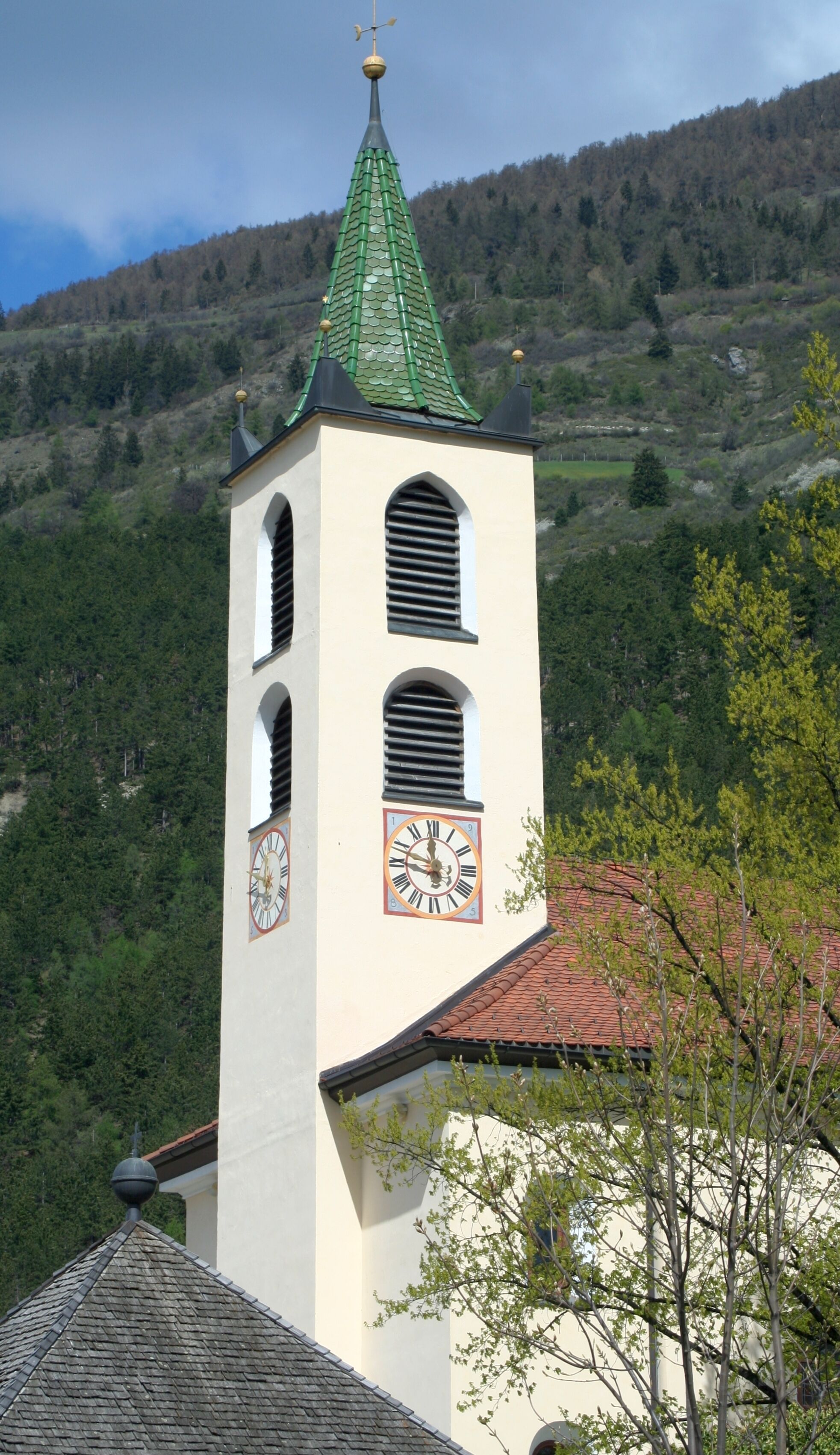A church tower with a clock, surrounded by a forested hillside.