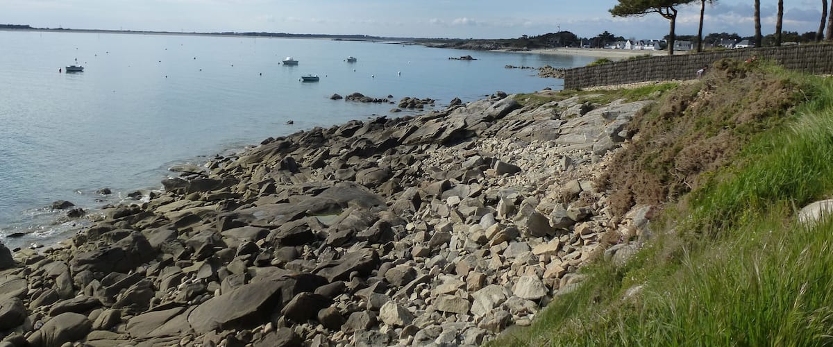 A rocky shoreline with calm water, boats, and a distant tree line.