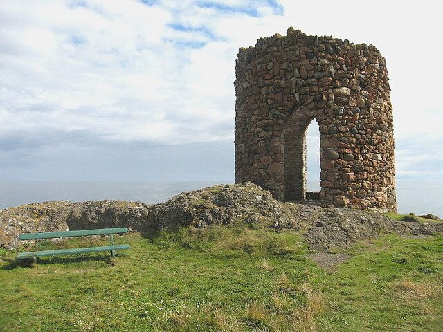 A stone archway structure on a grassy hill with a bench in the foreground.