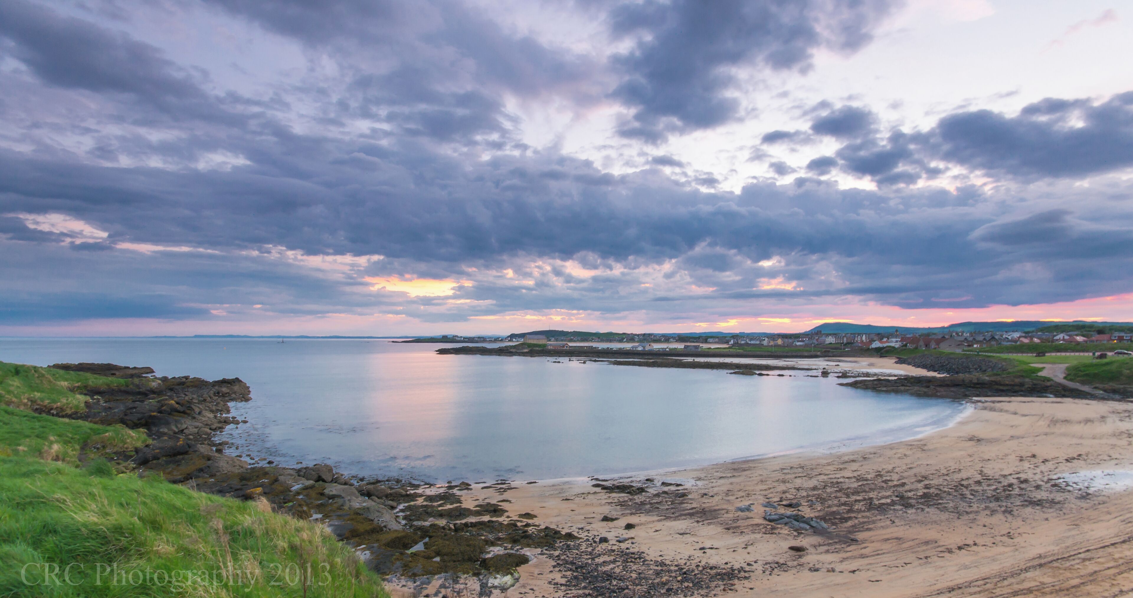 A coastal landscape with a sandy beach, rocky shoreline, and a sunset over the water.