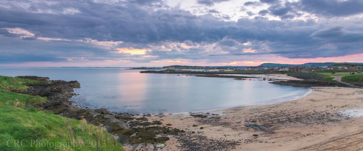 A coastal landscape with a sandy beach, rocky shoreline, and a sunset over the water.