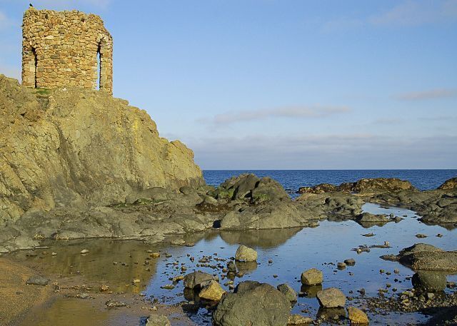 A coastal stone structure with a tower, surrounded by rocky terrain and calm waters.