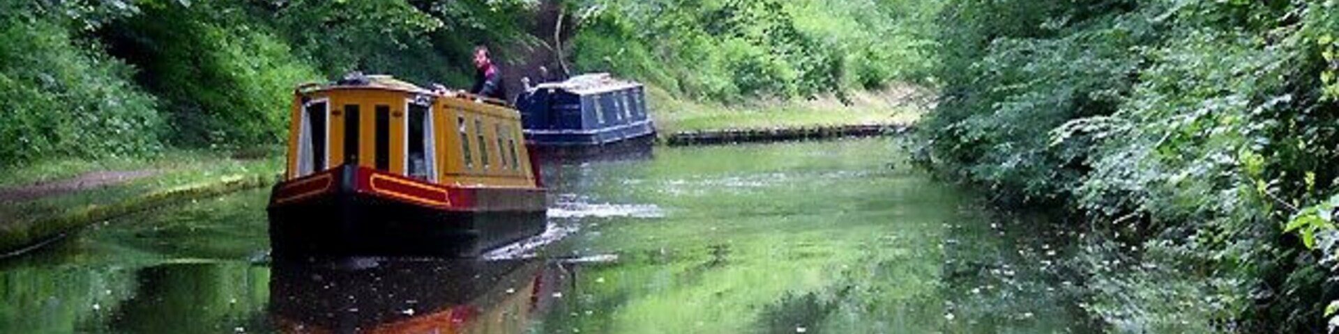 The Shropshire Union Canal north of Norbury, Staffordshire. An orange "Viking Afloat" hire boat is approaching having come through the Grub Street cutting. There are long, often straight sections of this canal which required either banking or cutting to avoid the need for locks. Although this was a more difficult procedure, the end route was shorter and the speed for horse drawn working boats could be maintained with (hopefully) no stoppages. The canal (formerly the Birmingham and Liverpool Junction Canal) was opened in 1835 at a time when railways were making serious inroads into the canal companies' businesses.