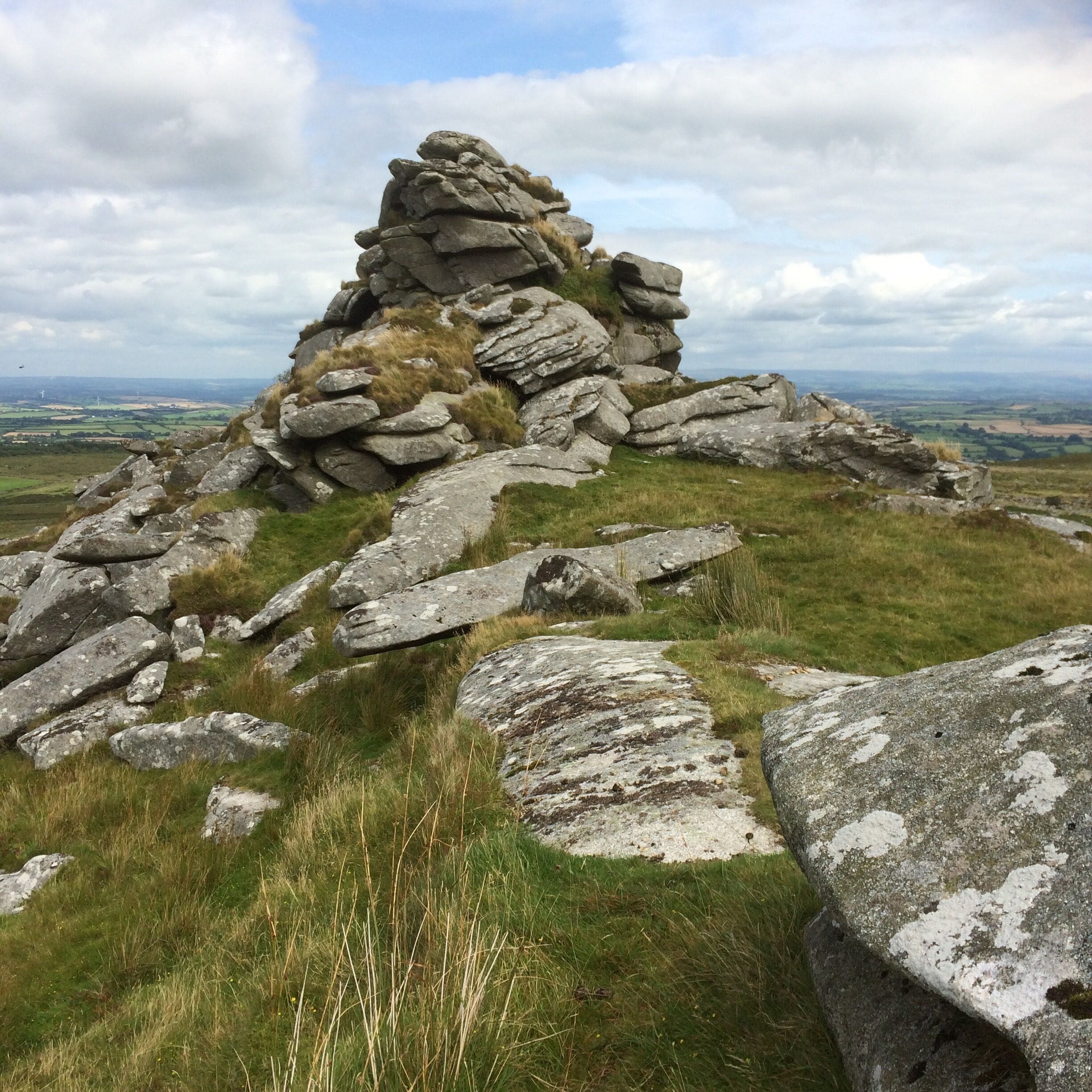 A rocky hill with a cairn on top, surrounded by grass and a distant view of the landscape.