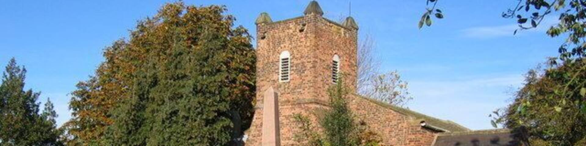 A historic stone church with a bell tower, surrounded by lush greenery and a brick wall.