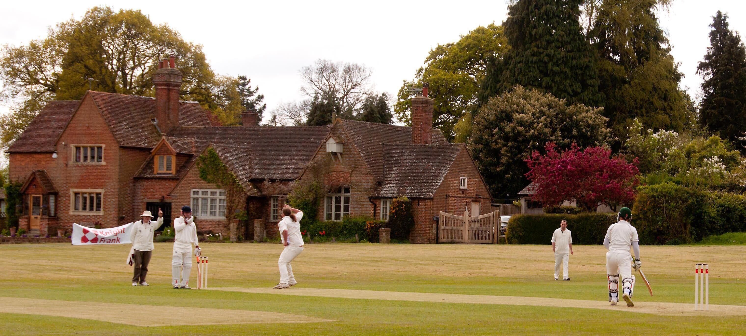 A cricket match in a grassy field with a brick building in the background.