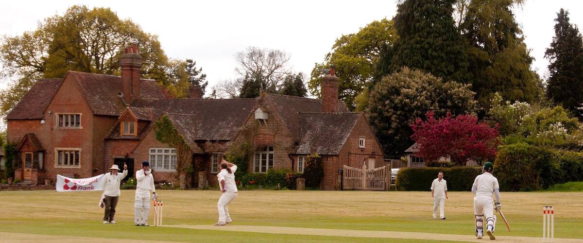 A cricket match in a grassy field with a brick building in the background.