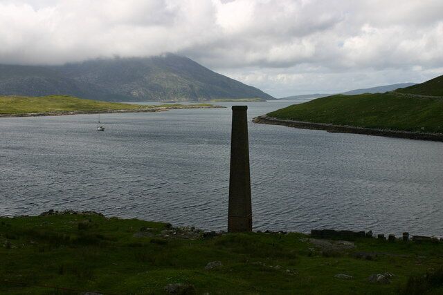 A body of water with a tall chimney-like structure in the foreground, surrounded by grassy hills and mountains in the background.