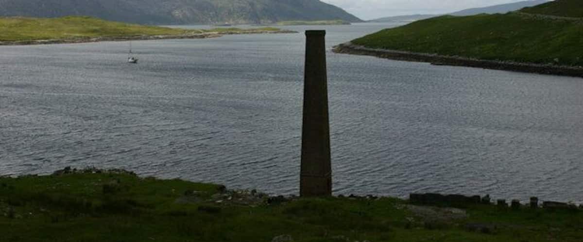 A body of water with a tall chimney-like structure in the foreground, surrounded by grassy hills and mountains in the background.