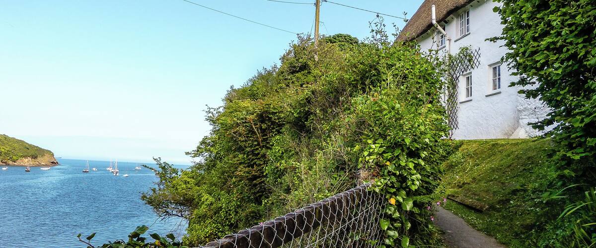 A coastal path with a white house, a thatched roof, and a view of the sea with boats.