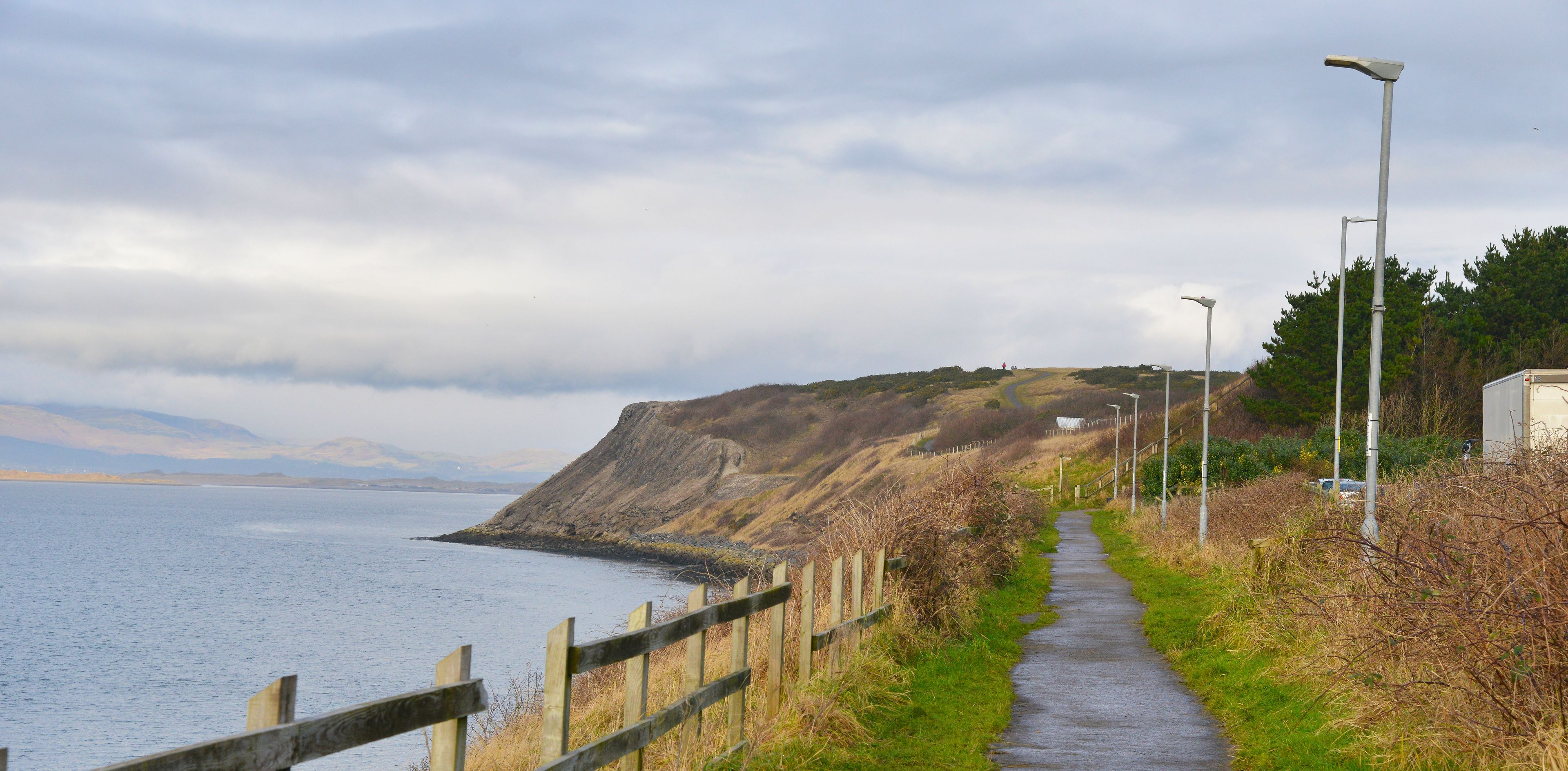 A coastal pathway with a wooden fence, street lamps, and a view of the sea and distant mountains.