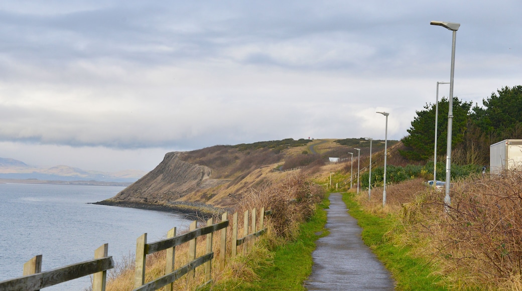 A coastal pathway with a wooden fence, street lamps, and a view of the sea and distant mountains.