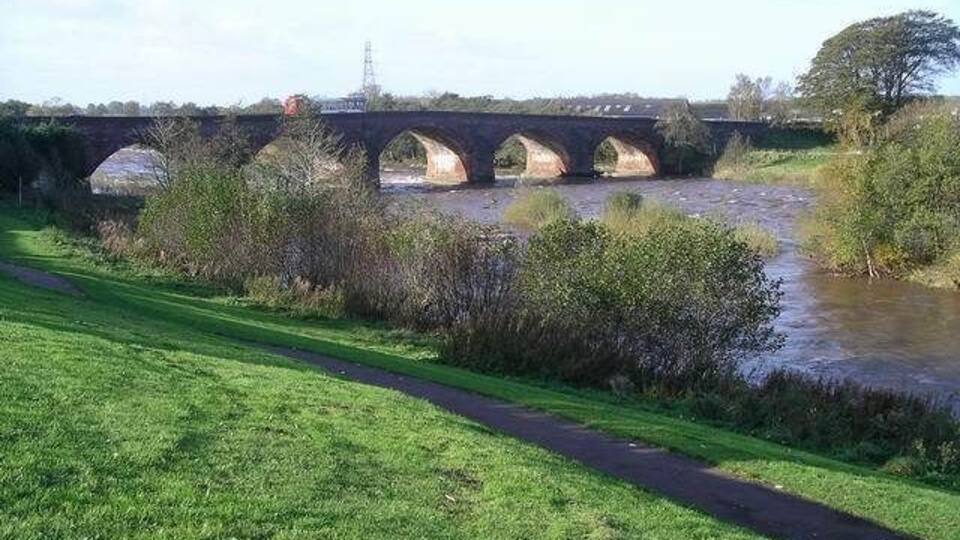 A stone bridge over a river, surrounded by greenery and a grassy path.