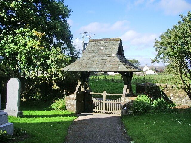 A stone gazebo with a pointed roof in a grassy area with a stone wall and a pathway leading to it.
