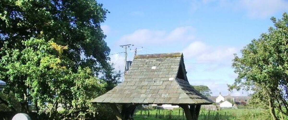 A stone gazebo with a pointed roof in a grassy area with a stone wall and a pathway leading to it.