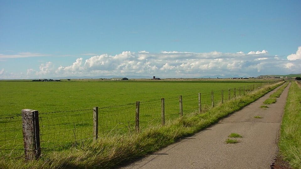 A rural road bordered by a green field and a fence, leading towards distant hills under a clear sky.
