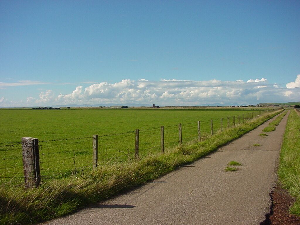 A rural road bordered by a green field and a fence, leading towards distant hills under a clear sky.