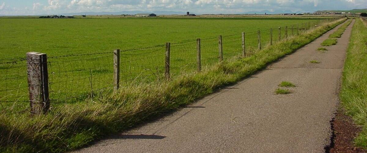 A rural road bordered by a green field and a fence, leading towards distant hills under a clear sky.