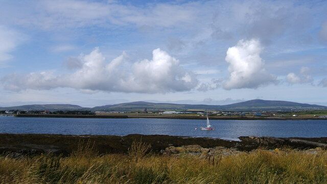 A coastal landscape with a body of water, a sailboat, and distant hills.