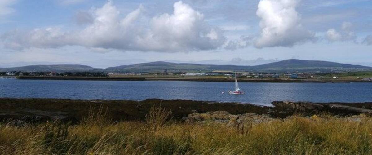 A coastal landscape with a body of water, a sailboat, and distant hills.