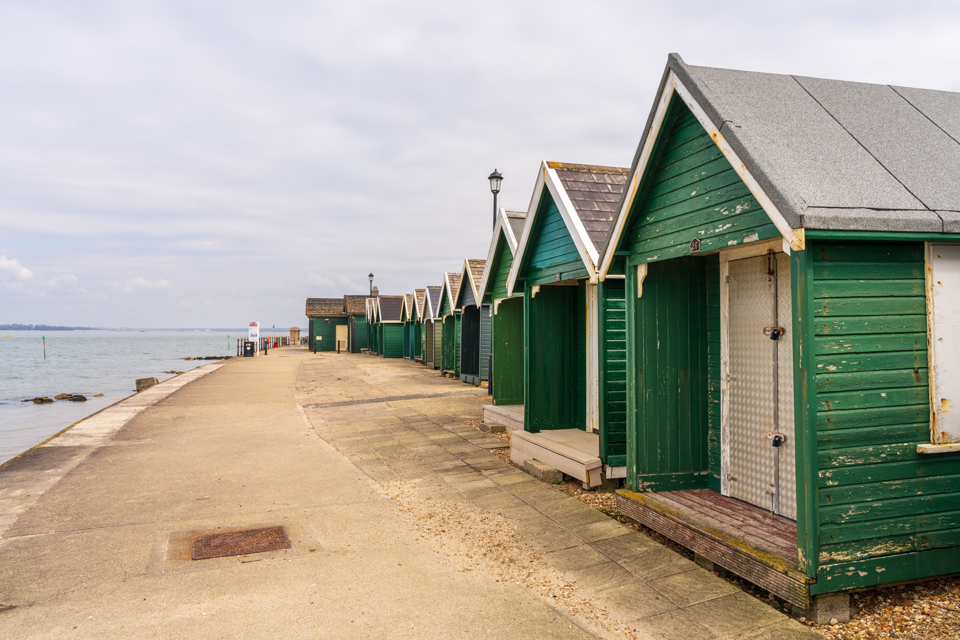 A row of green beach huts with a sandy walkway beside a body of water.