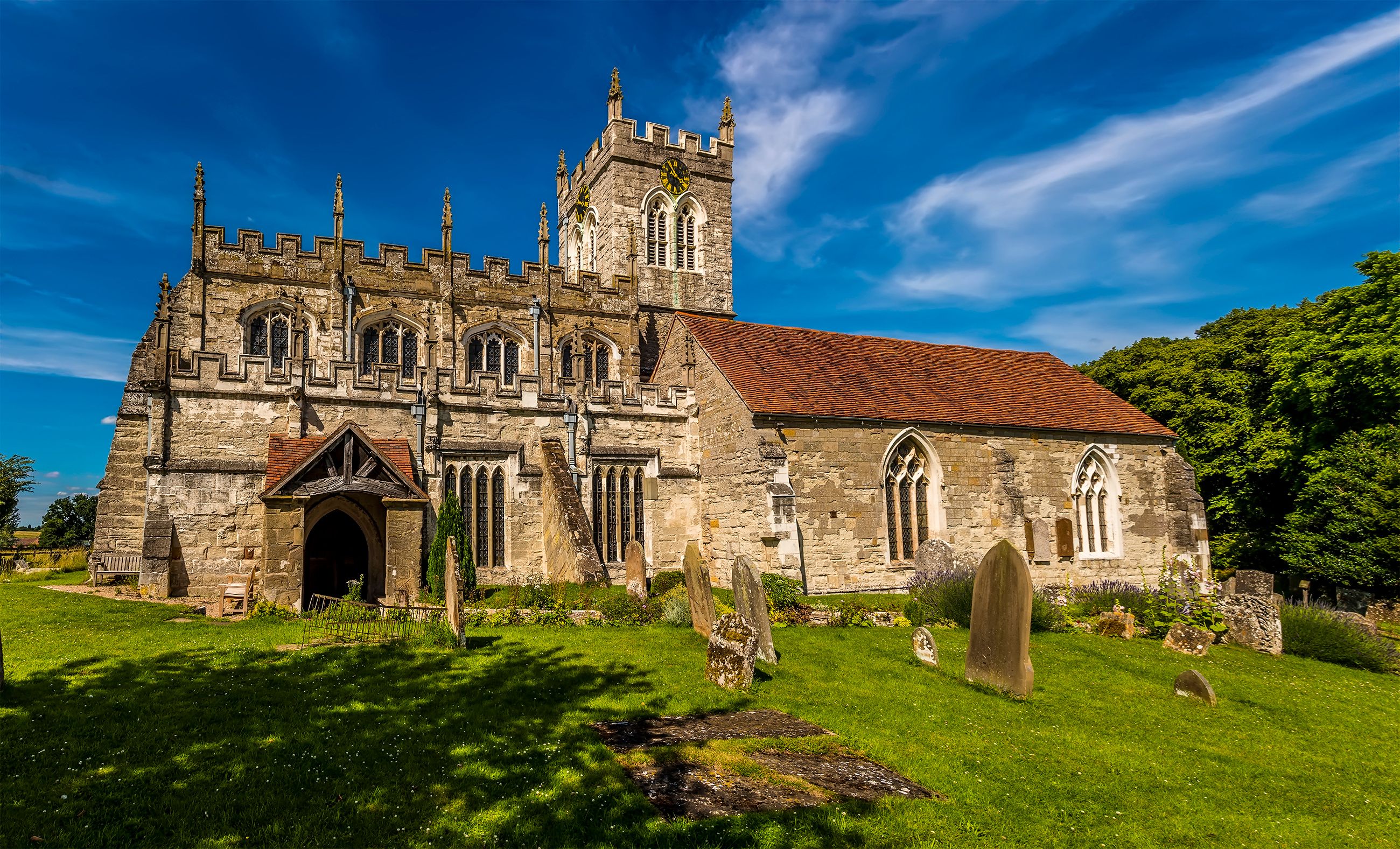 A historic stone church with a red-tiled roof, a tall bell tower, and a graveyard with headstones.