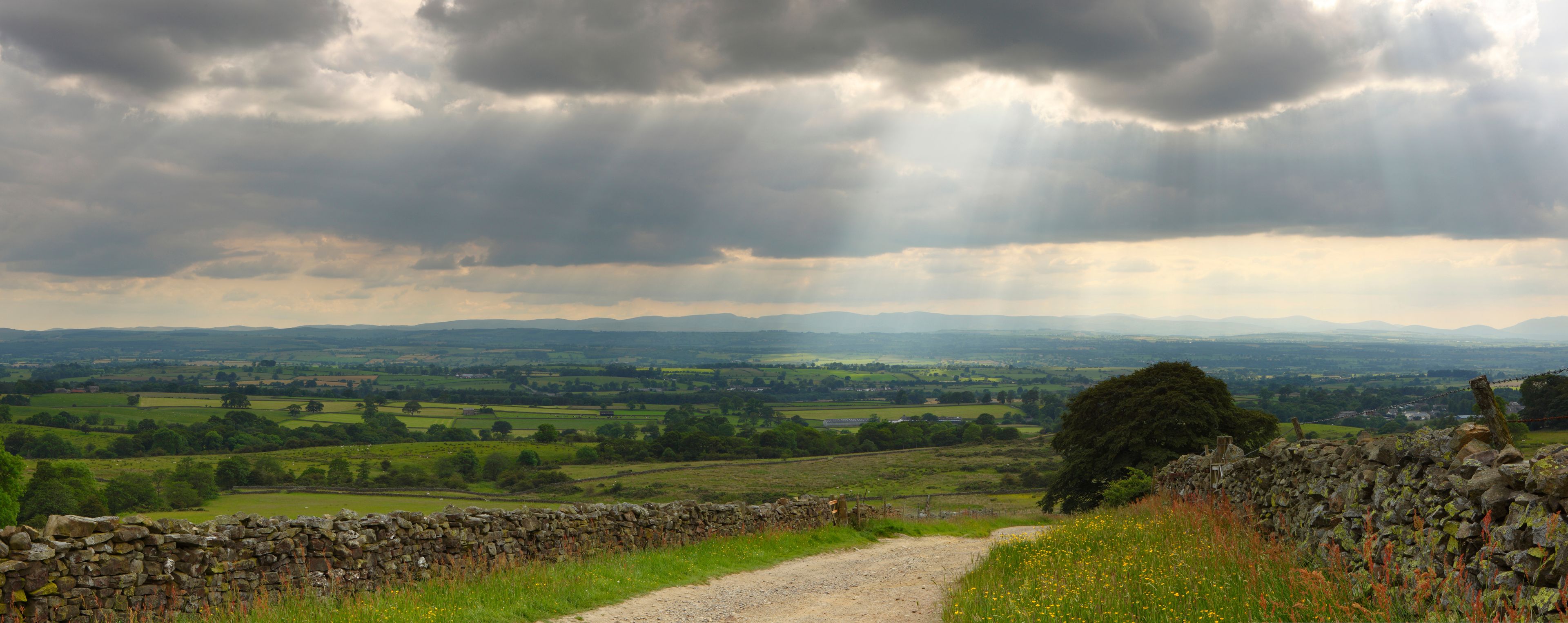 A rural landscape with a stone wall, a dirt path, and rolling hills in the distance under a cloudy sky.