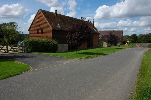A rural road leading to a wooden building with a sloped roof, surrounded by green grass and trees.