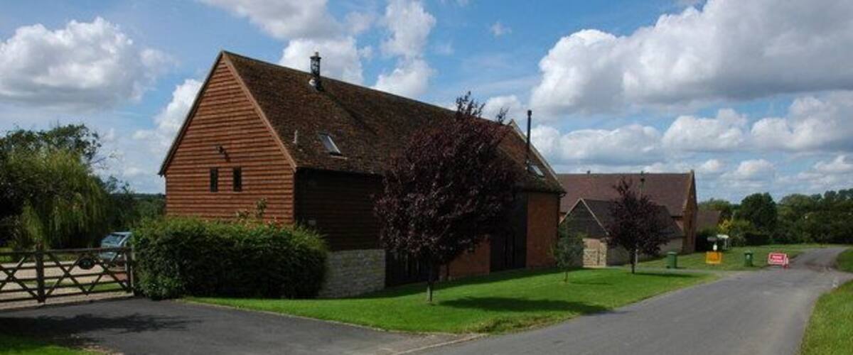 A rural road leading to a wooden building with a sloped roof, surrounded by green grass and trees.