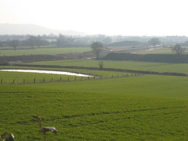 view of Gloucester SW bypass under construction. Robinswood Hill on horizon. Gloucester Sharpness Canal at field boundary beyond construction line. Water in foreground is a temporary pond.