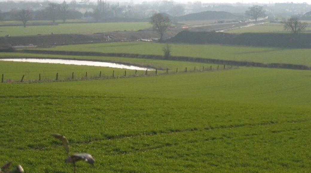 view of Gloucester SW bypass under construction. Robinswood Hill on horizon. Gloucester Sharpness Canal at field boundary beyond construction line. Water in foreground is a temporary pond.