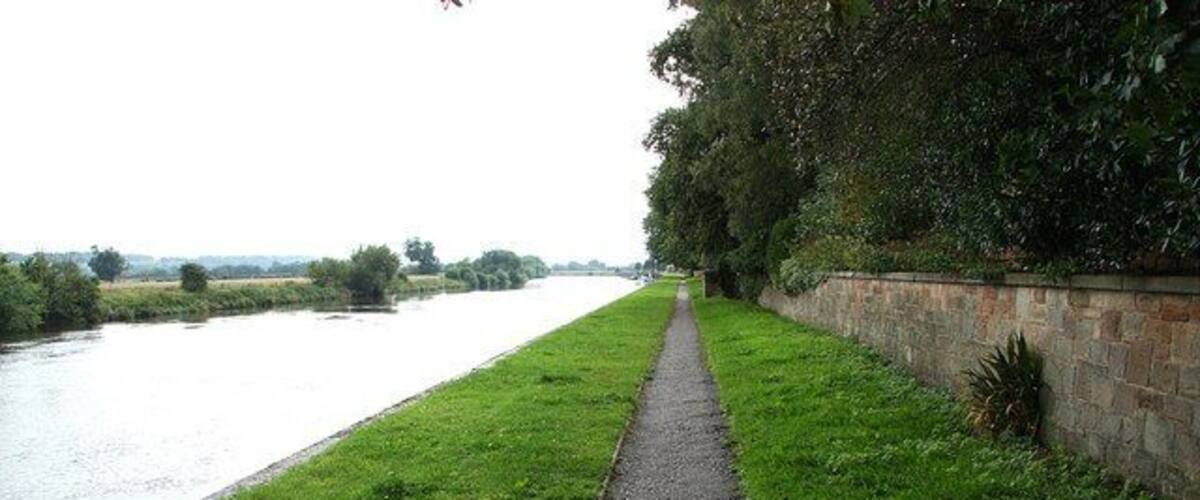 A narrow path beside a river, bordered by grass and a stone wall.