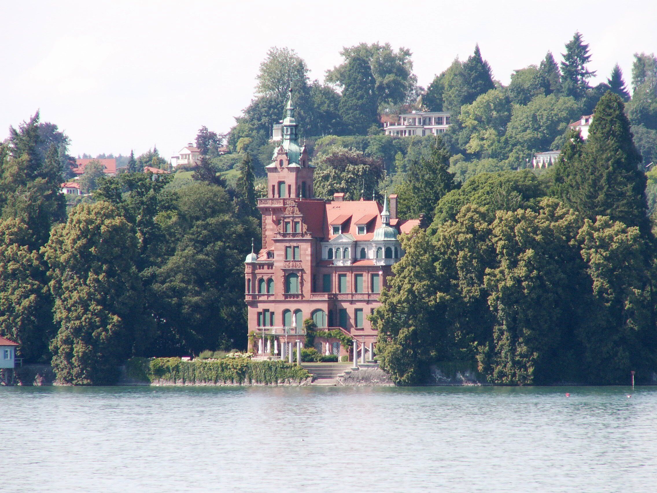 A large, ornate building with a tower, surrounded by dense trees and located near a body of water.