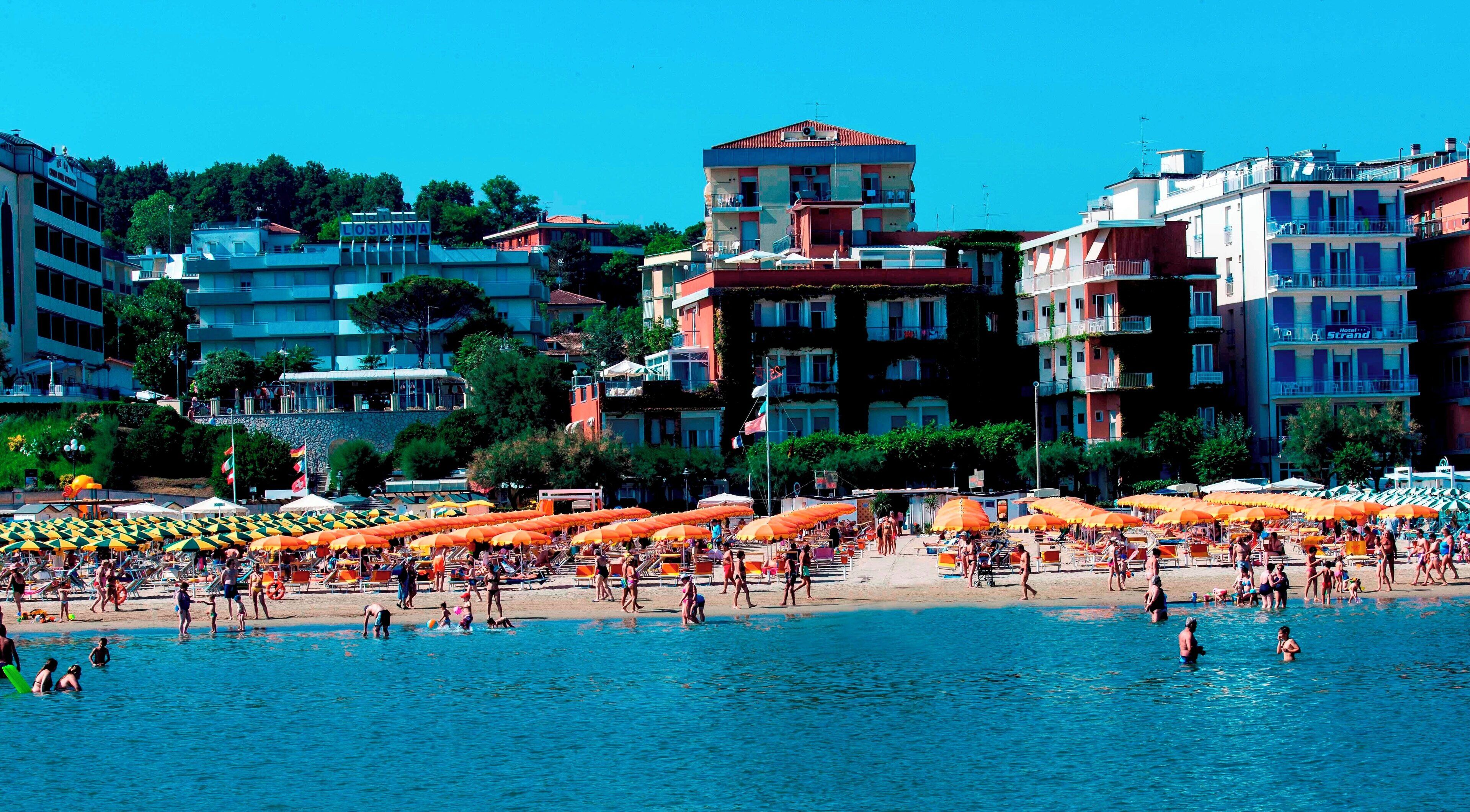 A crowded beach with orange umbrellas and people swimming.