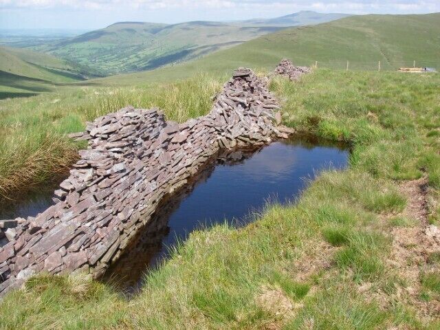 Dry stone wall or wet stone wall? The dilapidated wall which runs across Fan Fraith dips its feet in a pool. Pen y Fan, Fan Frynach and the north end of Fan Nedd are seen in the distance. This pool is but one of many (mostly) alongside the wall from which presumably the stones for its construction were unearthed.