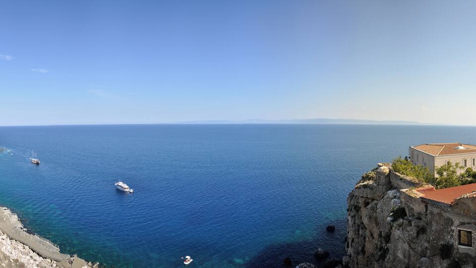 A coastal landscape with a clear blue sea, rocky cliffs, and a historic building with a red-tiled roof.
