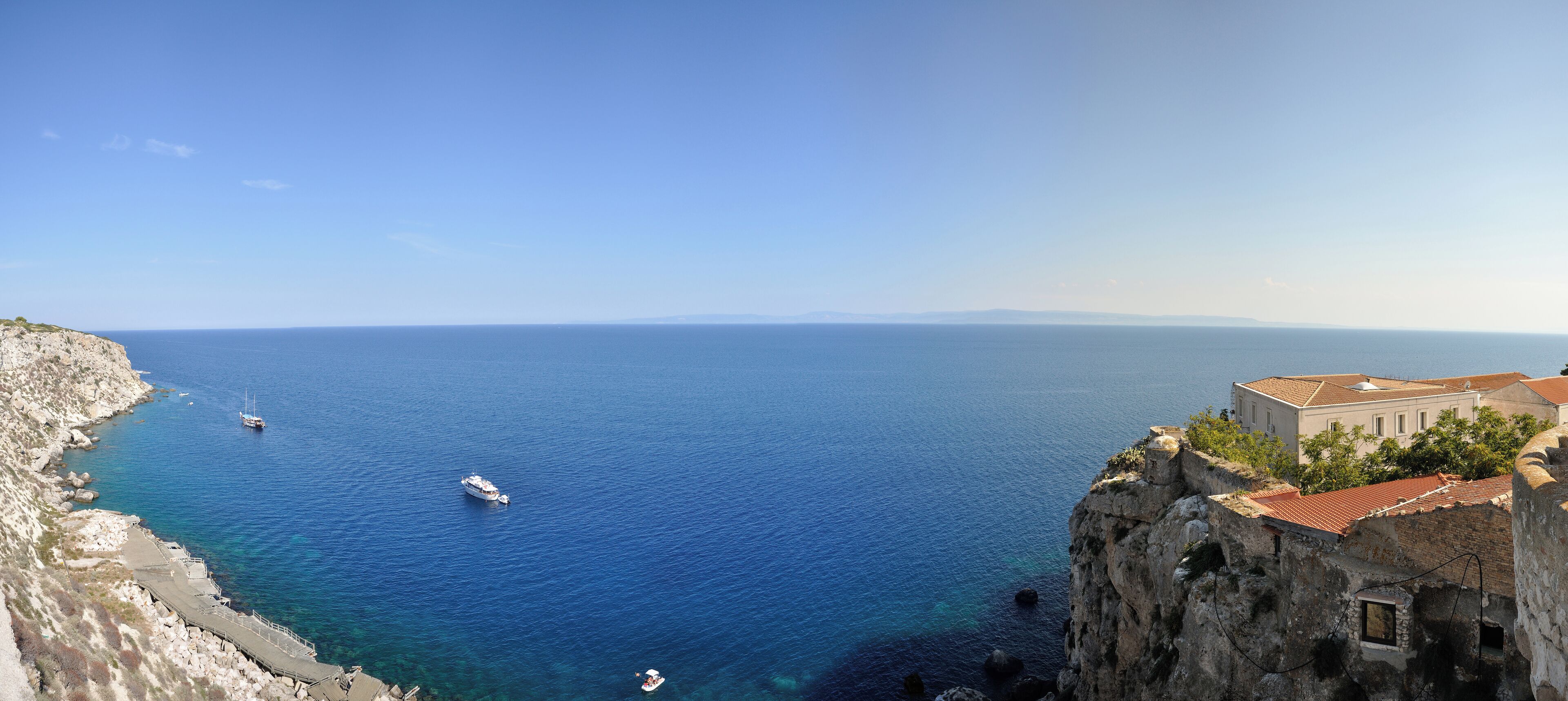 A coastal view with a clear blue sea, rocky cliffs, and a historic building with a red-tiled roof.