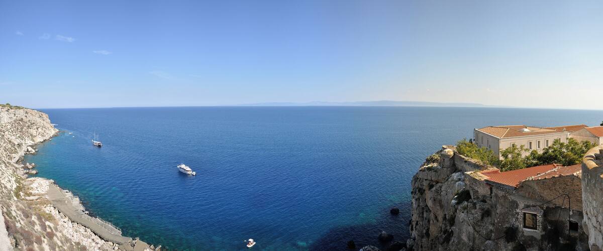 A coastal view with a clear blue sea, rocky cliffs, and a historic building with a red-tiled roof.
