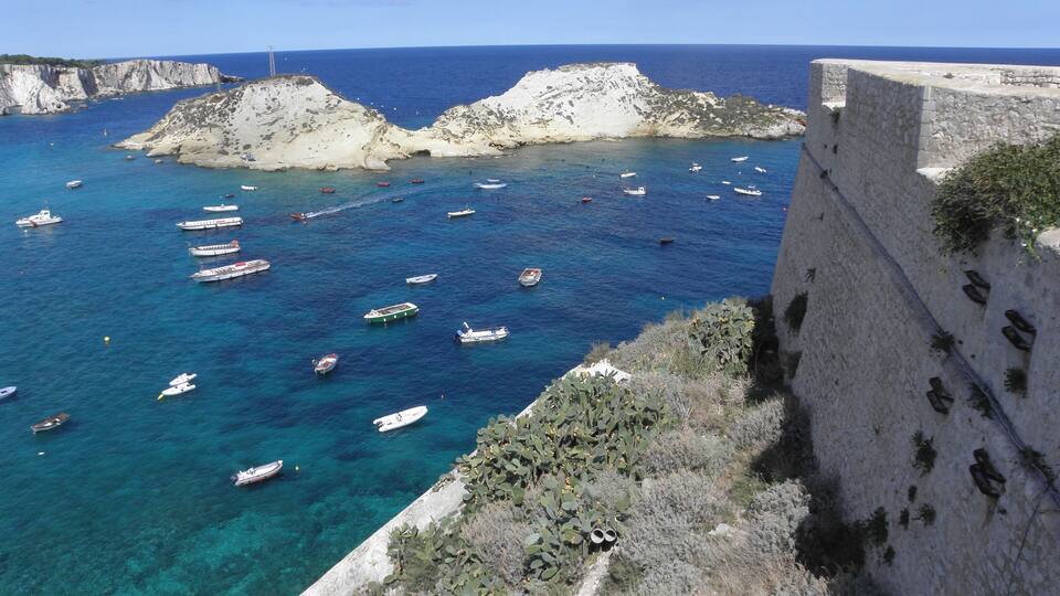 A coastal landscape with clear blue waters, rocky cliffs, and a stone wall.