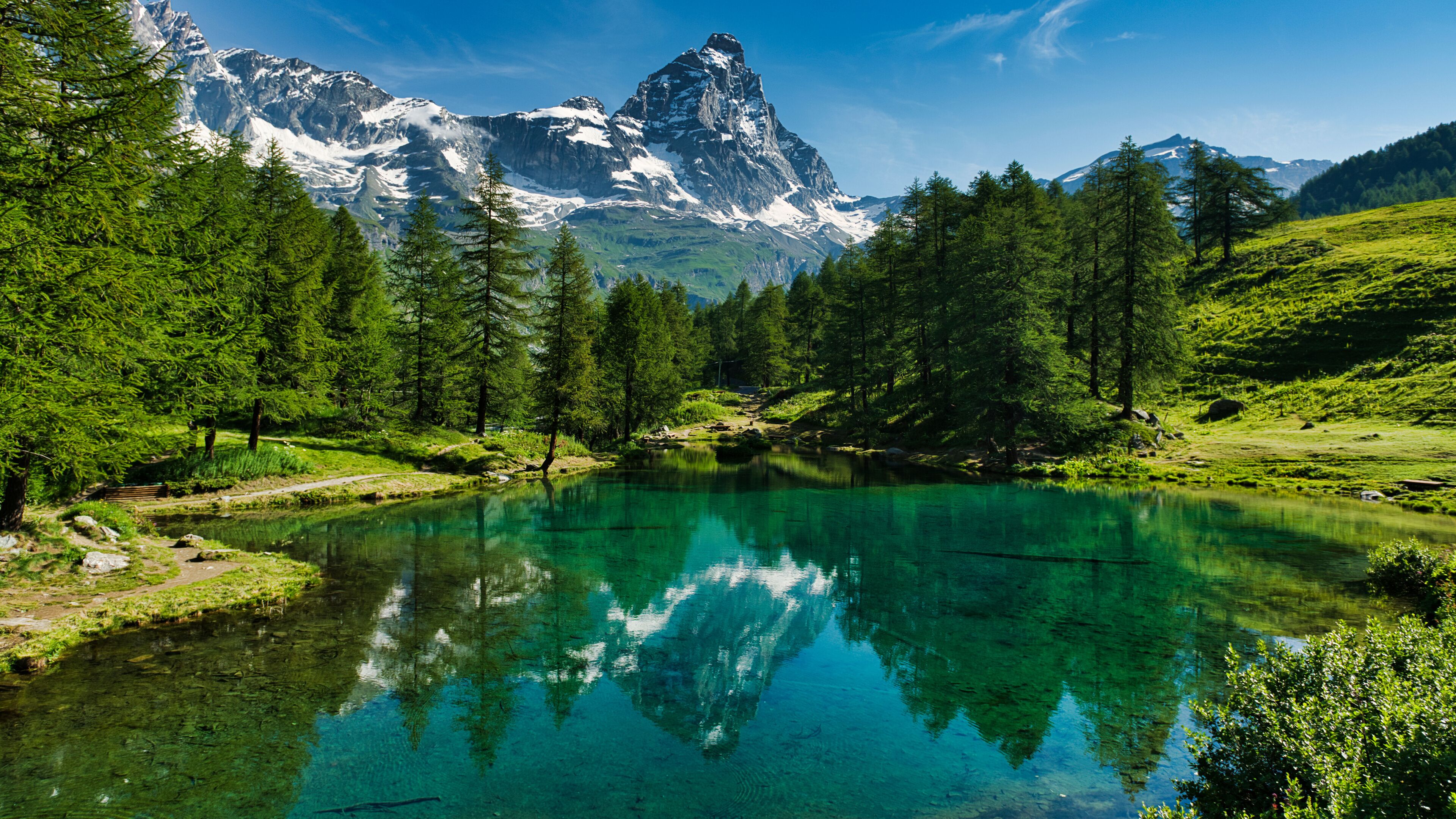 A clear mountain lake surrounded by dense forest and snow-capped peaks.