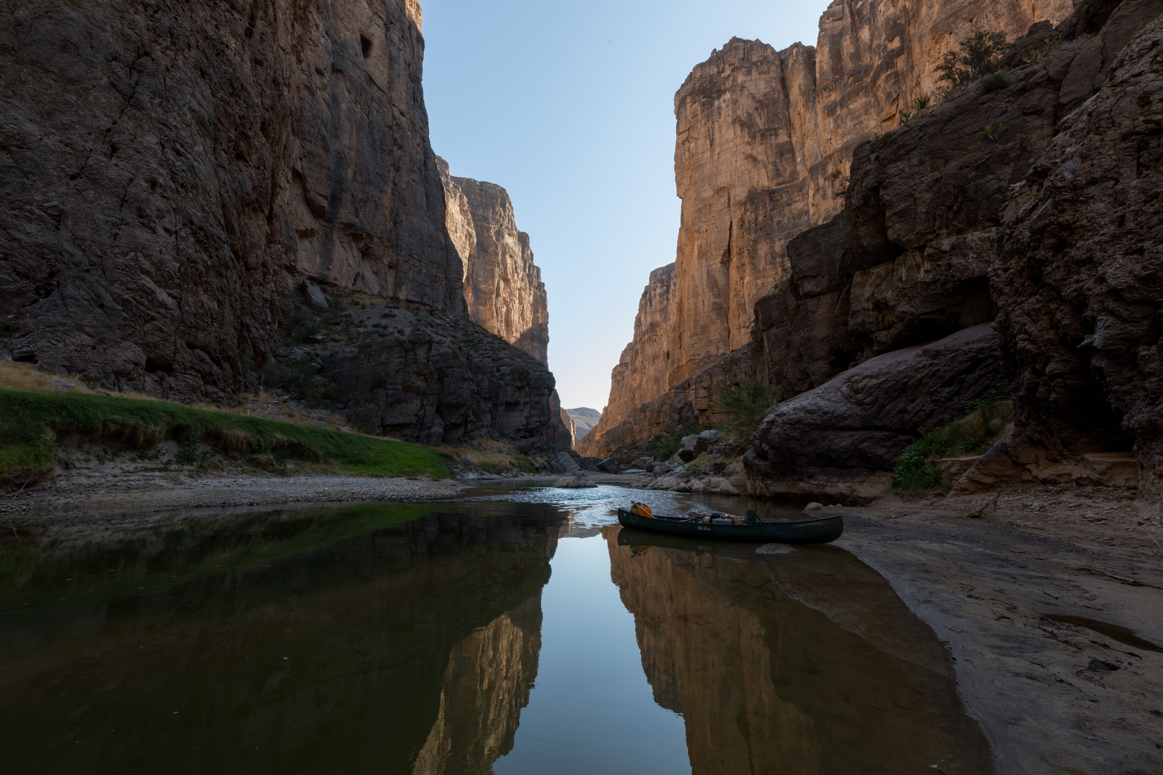 A narrow canyon with steep rock walls and a calm river reflecting the scenery.