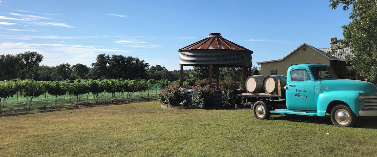 A vintage turquoise truck parked in a grassy field with a metal gazebo labeled "GUTIER" and a building labeled "Fiesta Winery" in the background.