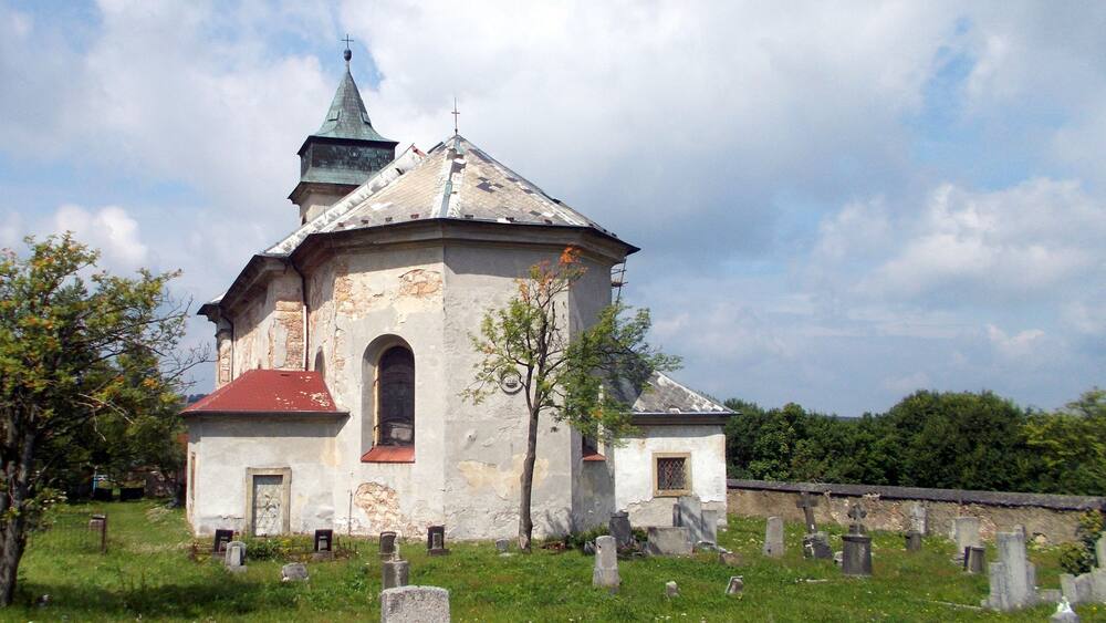 Une vieille église avec un toit rouge et une flèche verte, entourée d’un cimetière.