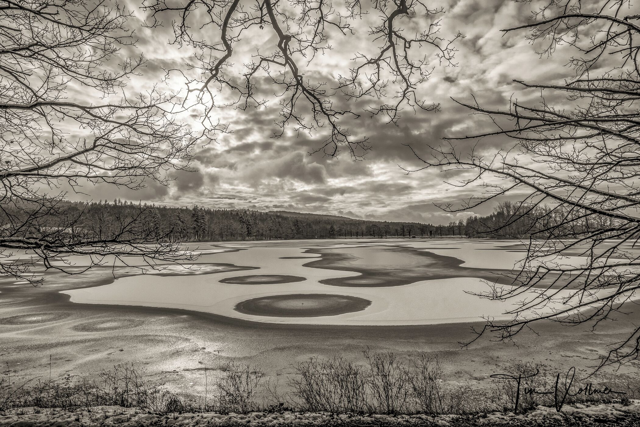 A frozen lake with circular patterns, surrounded by trees and a cloudy sky.