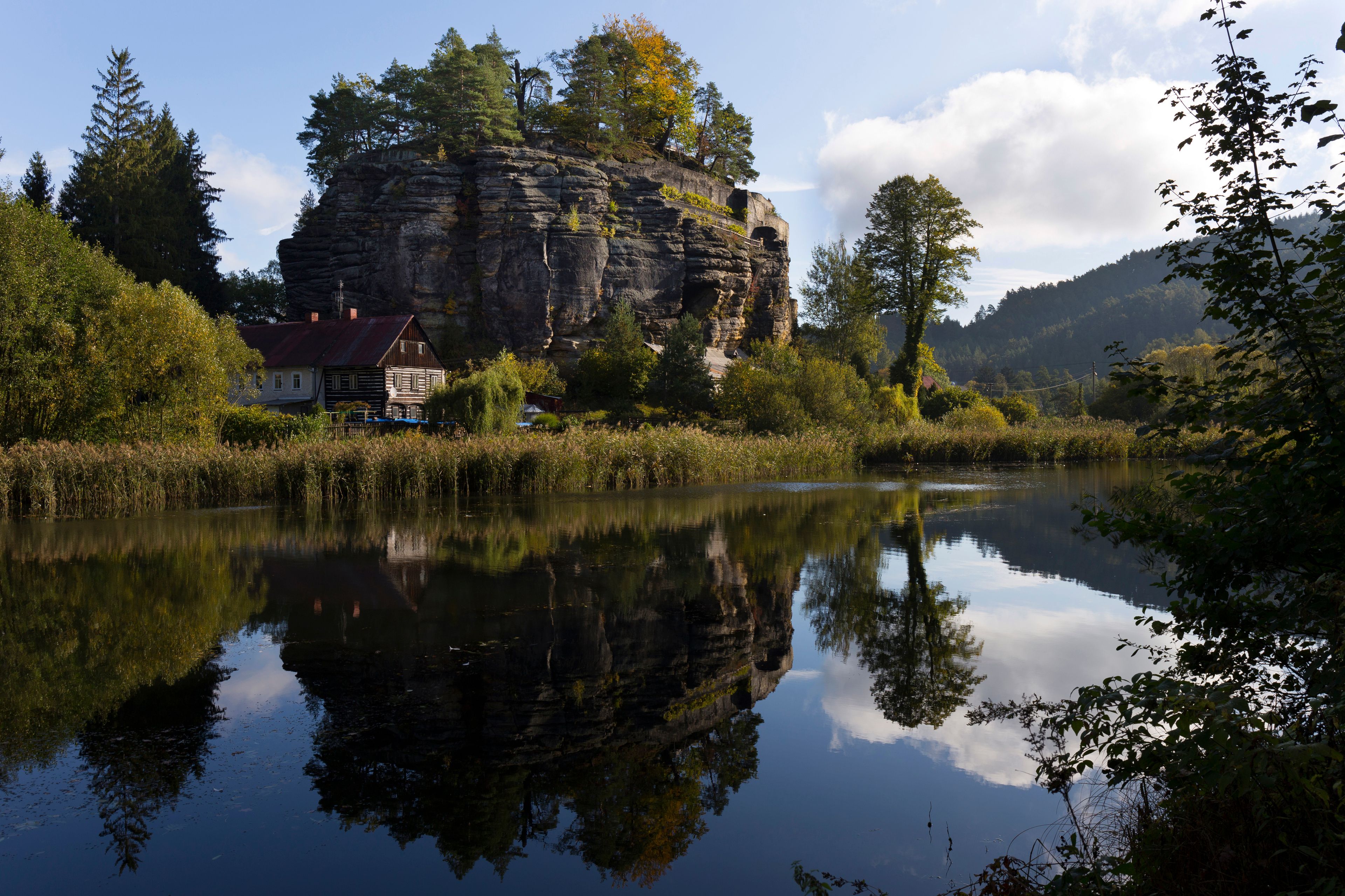 A calm river reflecting a rocky cliff and a traditional house with a red roof.