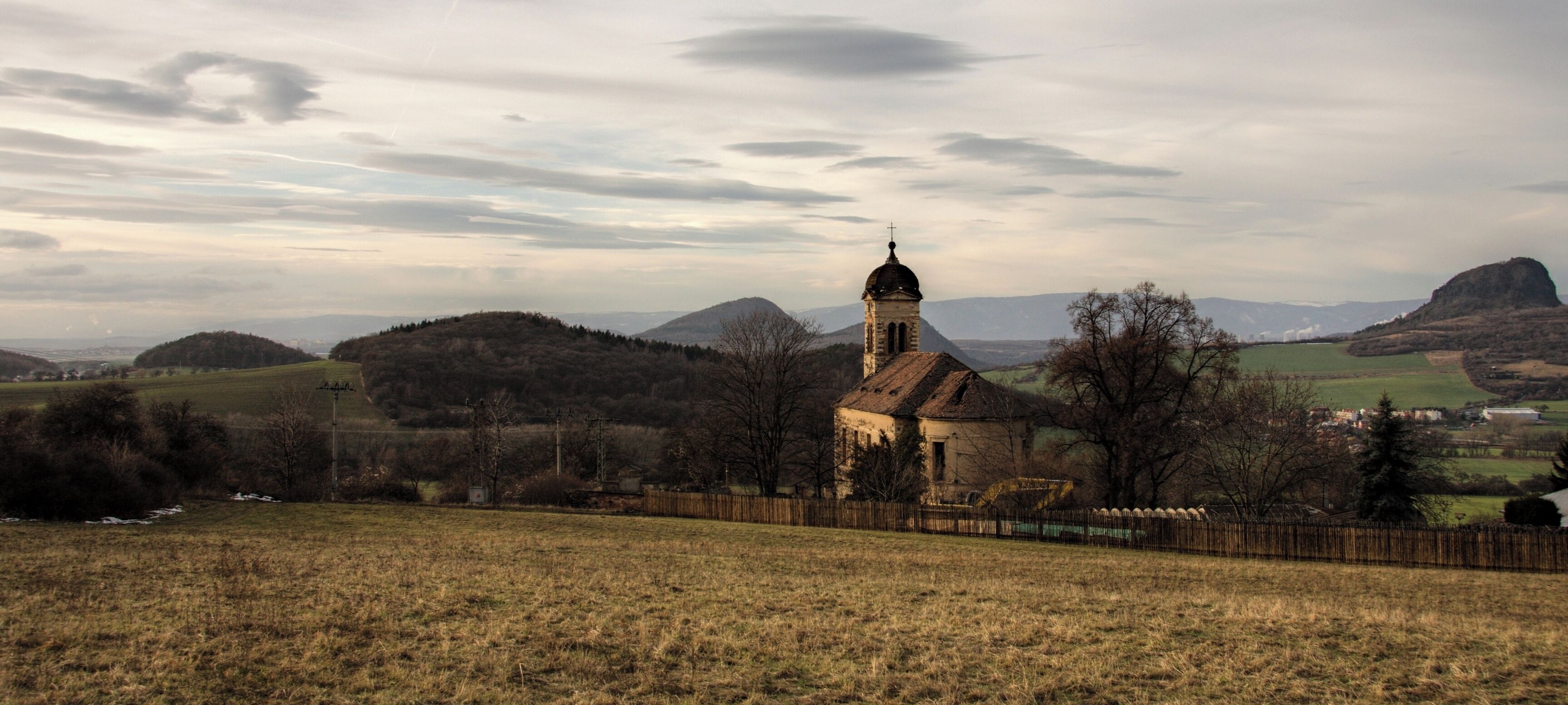 A church with a bell tower, surrounded by rolling hills and a cloudy sky.